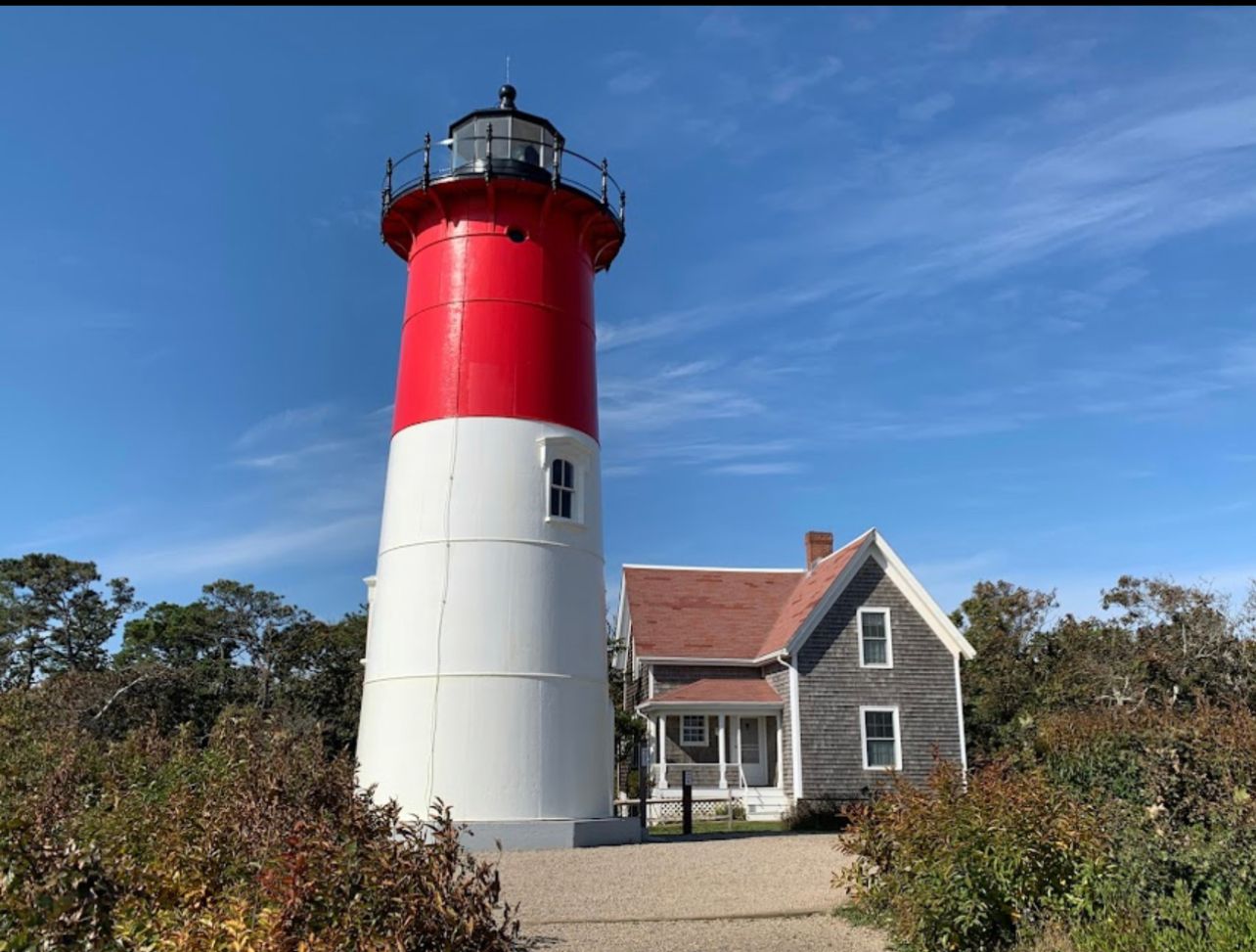 A tall lighthouse with a white base and red top stands next to a gray house with a red roof under a clear blue sky.