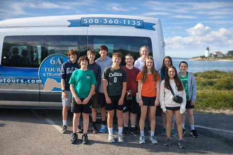 A group of eleven people stands in front of a 6A Tours van near a coastal lighthouse.