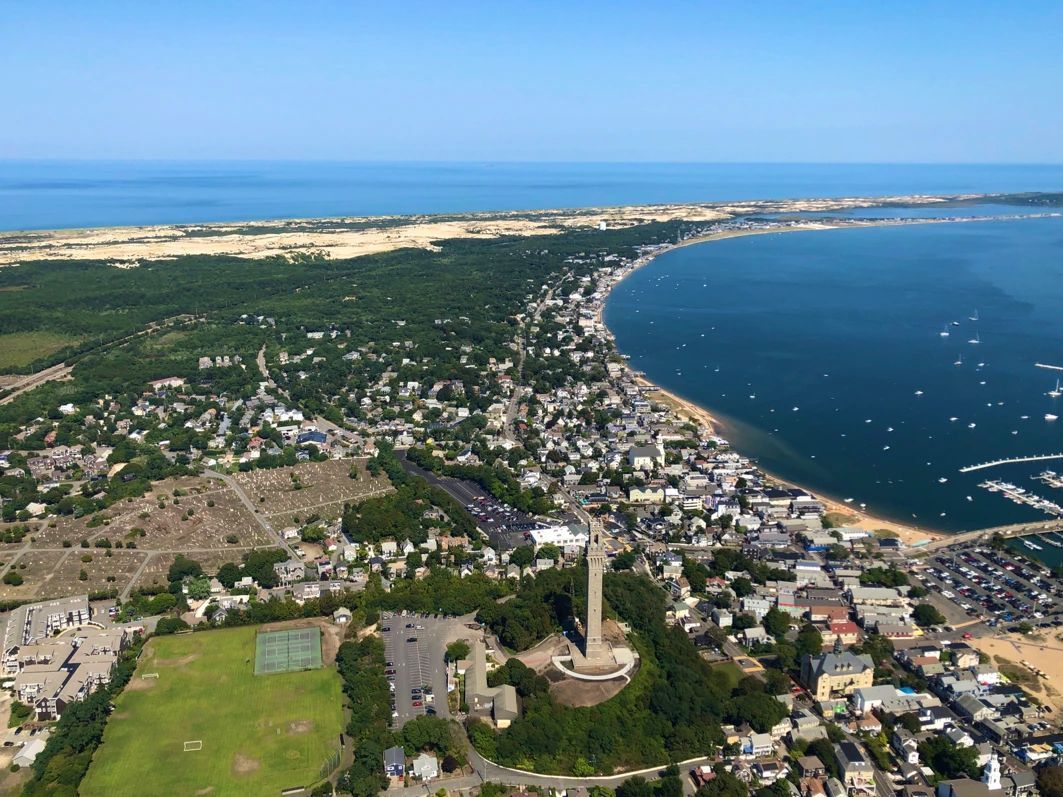 Aerial view of Provincetown, Massachusetts, with the Pilgrim Monument prominently displayed, coastline, and blue ocean.