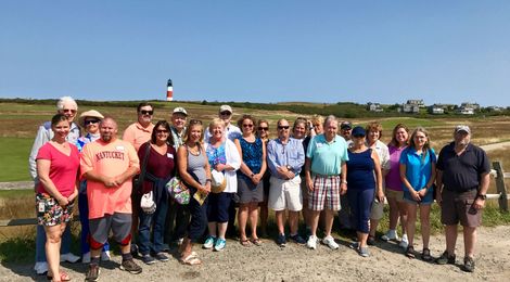 A large group stands together outdoors in a grassy field with a lighthouse in the background on a sunny day.