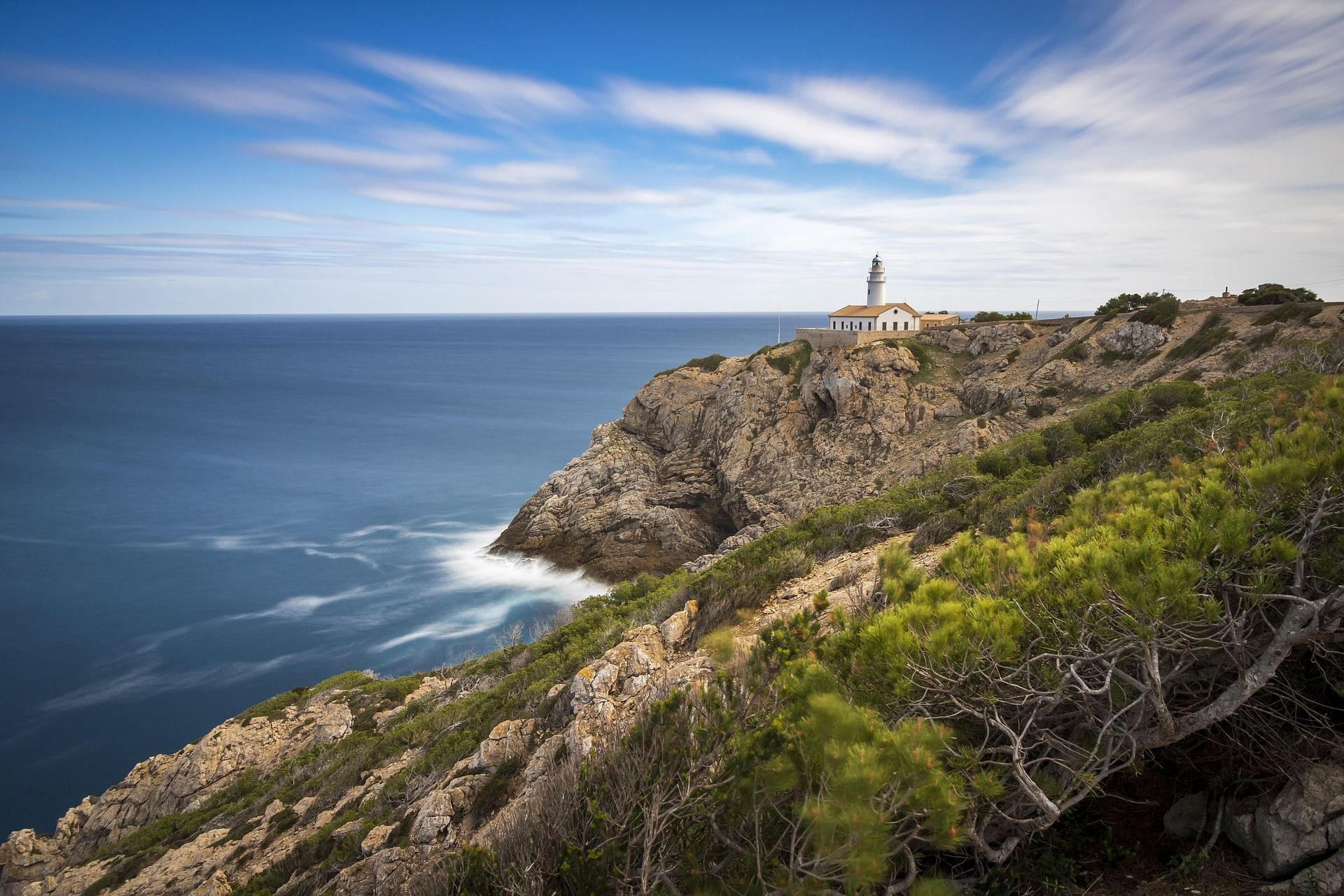 Lighthouse on a rocky cliff overlooking the ocean. Blue sky, white clouds, and green vegetation.