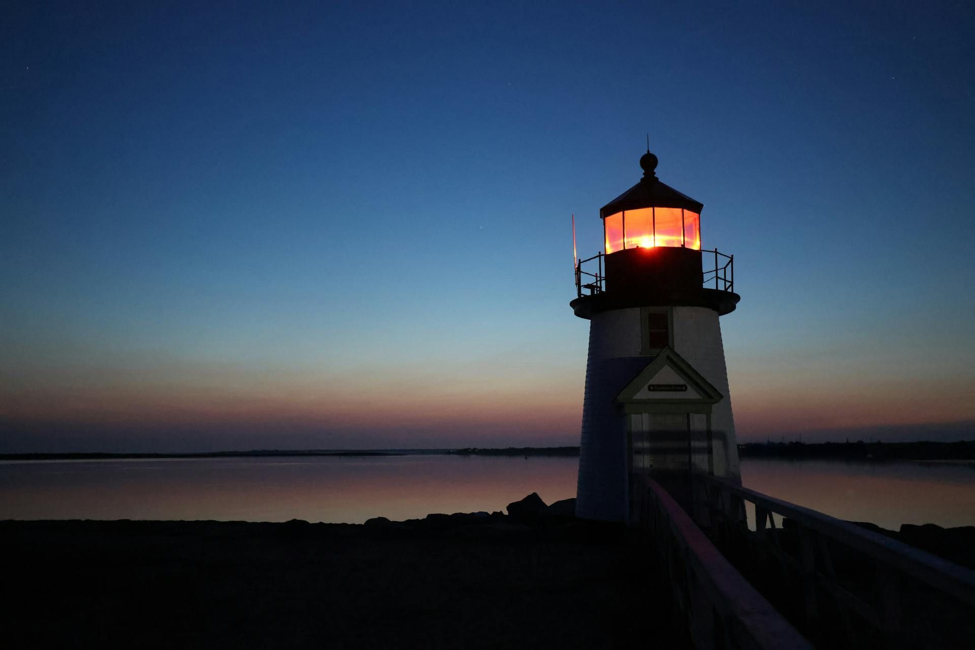 Lighthouse at dusk; light shining, silhouetted against a darkening sky and calm water.