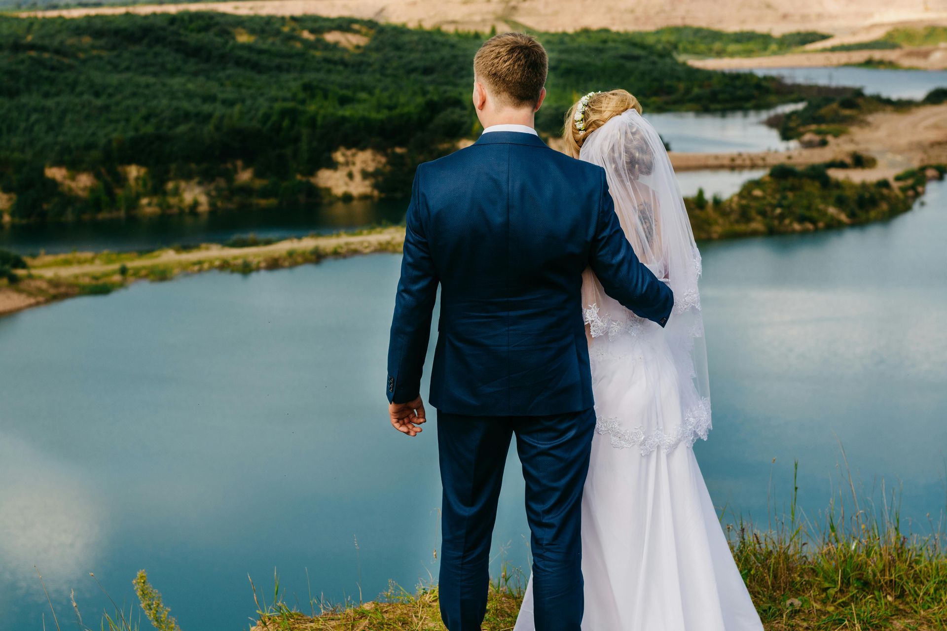 A bride and groom in wedding attire stand together, back to the camera, overlooking a scenic lake and hilly landscape.