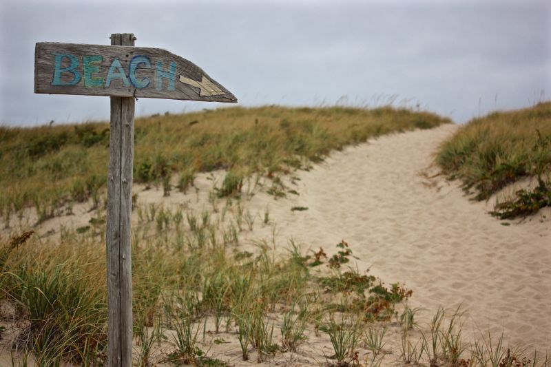 A rustic wooden sign pointing toward a sandy path leading through dunes covered in beach grass under a cloudy sky.