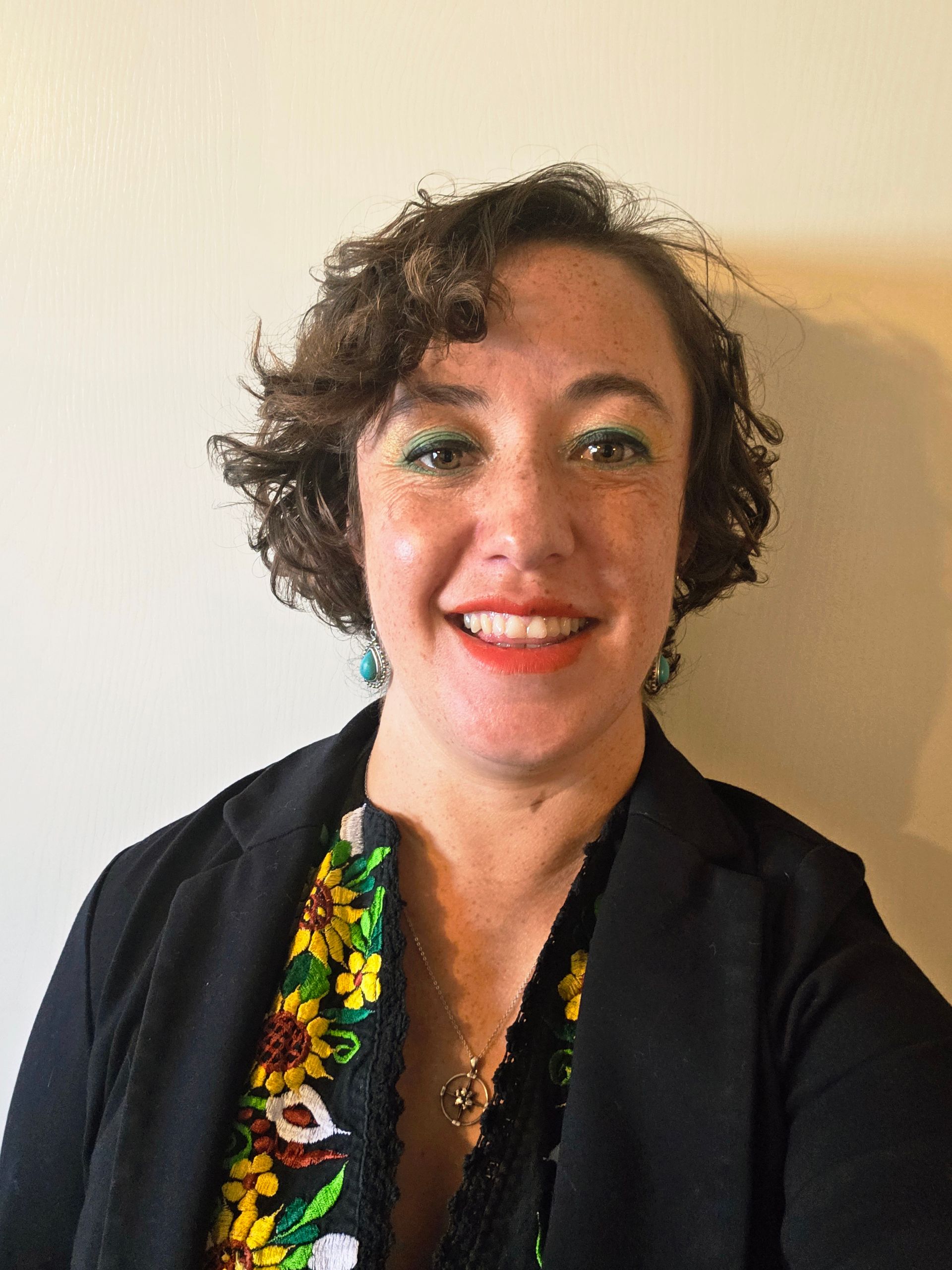 Professional headshot of Amber Miland, a smiling woman with short curly brown hair, green eye makeup, and red lipstick, wearing turquoise earrings, a black blazer, and a black embroidered blouse with yellow and green floral patterns, posed against a neutral background.