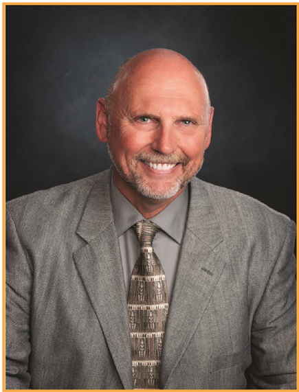 Professional headshot of Eric Smith, a smiling bald man with a neatly trimmed beard, wearing a gray suit jacket, patterned tie, and light gray shirt, posed against a dark studio background.