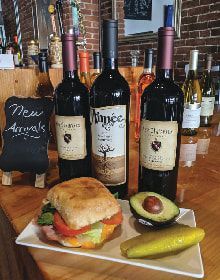 A sandwich , avocado , pickles and wine bottles on a table.
