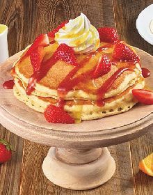 A stack of pancakes with strawberries and whipped cream on a wooden plate.