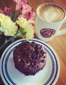 A chocolate cupcake is on a plate next to a cup of coffee and flowers.