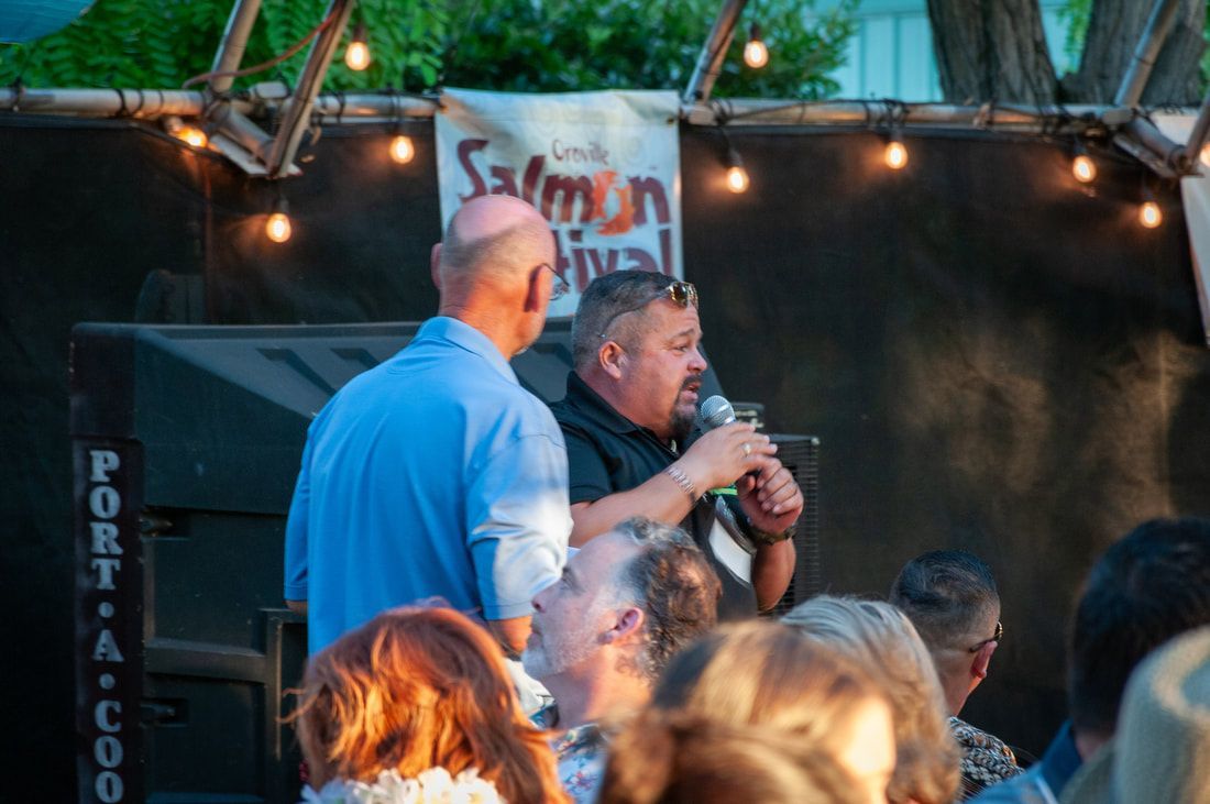 A man is speaking into a microphone in front of a crowd at a salmon festival.