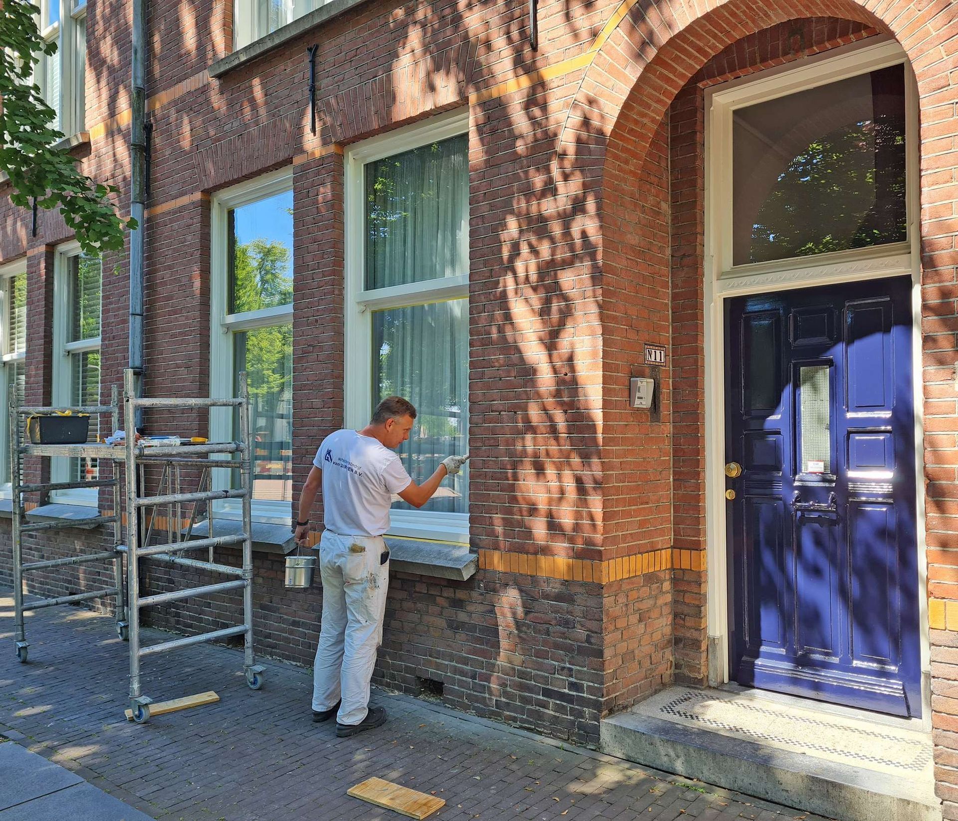 Man schildert raamkozijnen op een bakstenen gebouw, blauwe deur. Steiger, zonnige dag.
