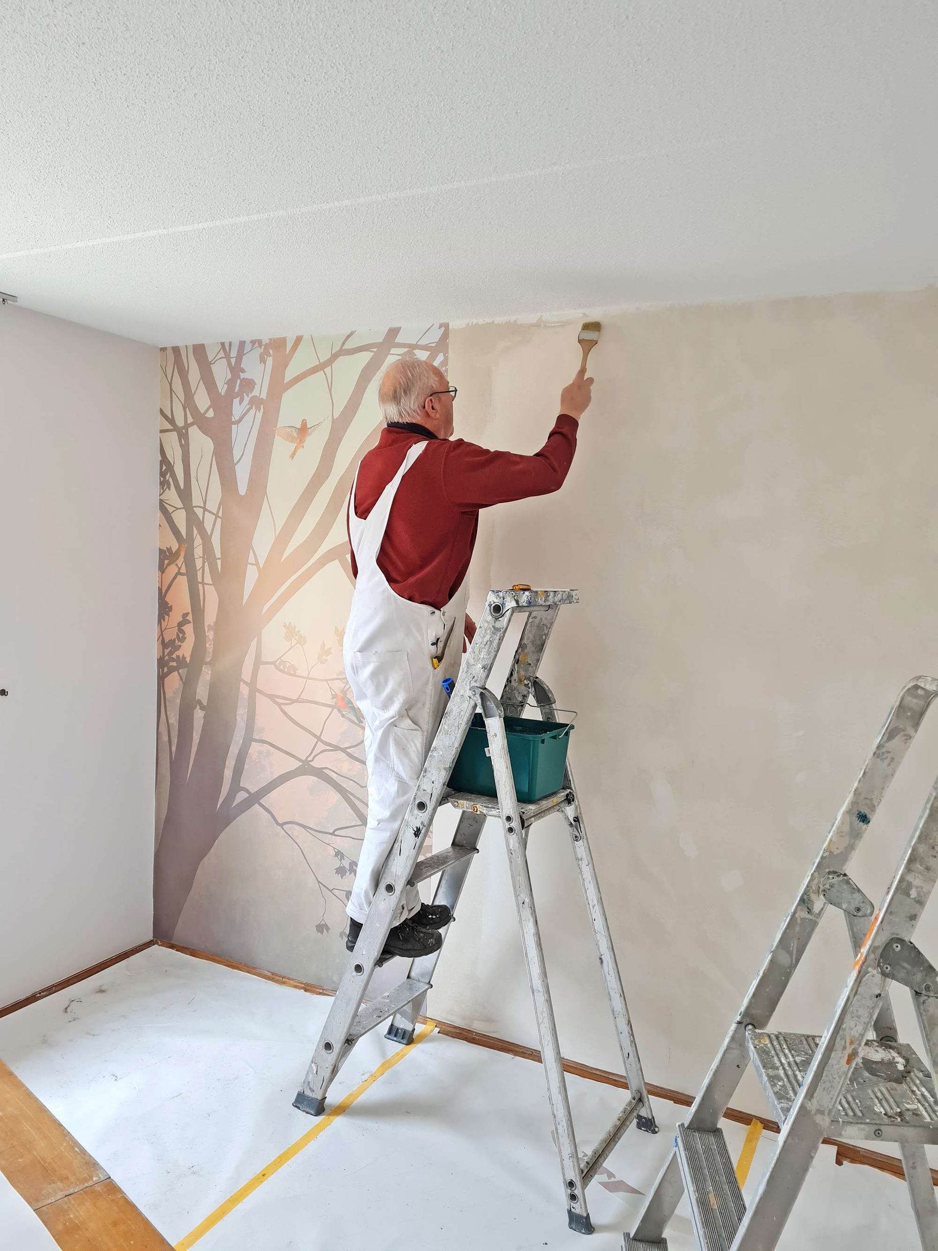 Man op een ladder schildert een muur met een kwast; op de muur ernaast is een muurschildering van een boom te zien.