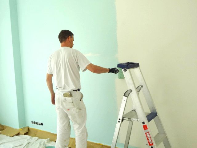 Schilder in witte kleding, met een roller, schildert een muur met lichtblauwe verf. Er is een ladder aanwezig.