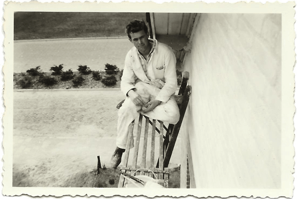 Man in witte overall op een ladder, die een witte muur schildert. Buiten, vlakbij een veld.