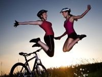 Two women are jumping in the air next to a bicycle.