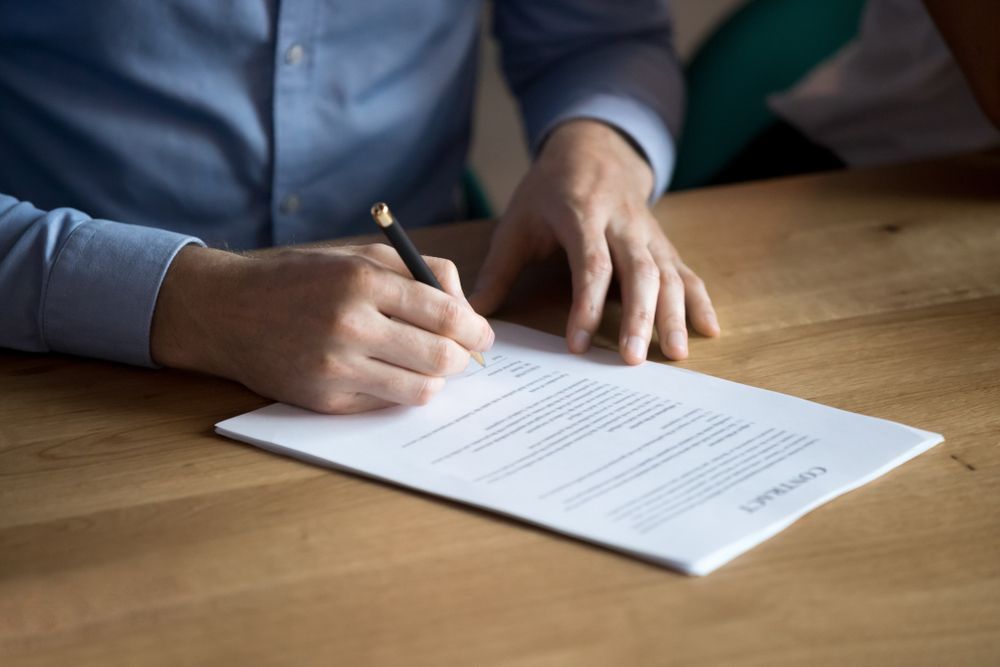 A Man is Signing a Document With a Pencil on a Wooden Table — MoneySmith Group In Kingscliff, NSW