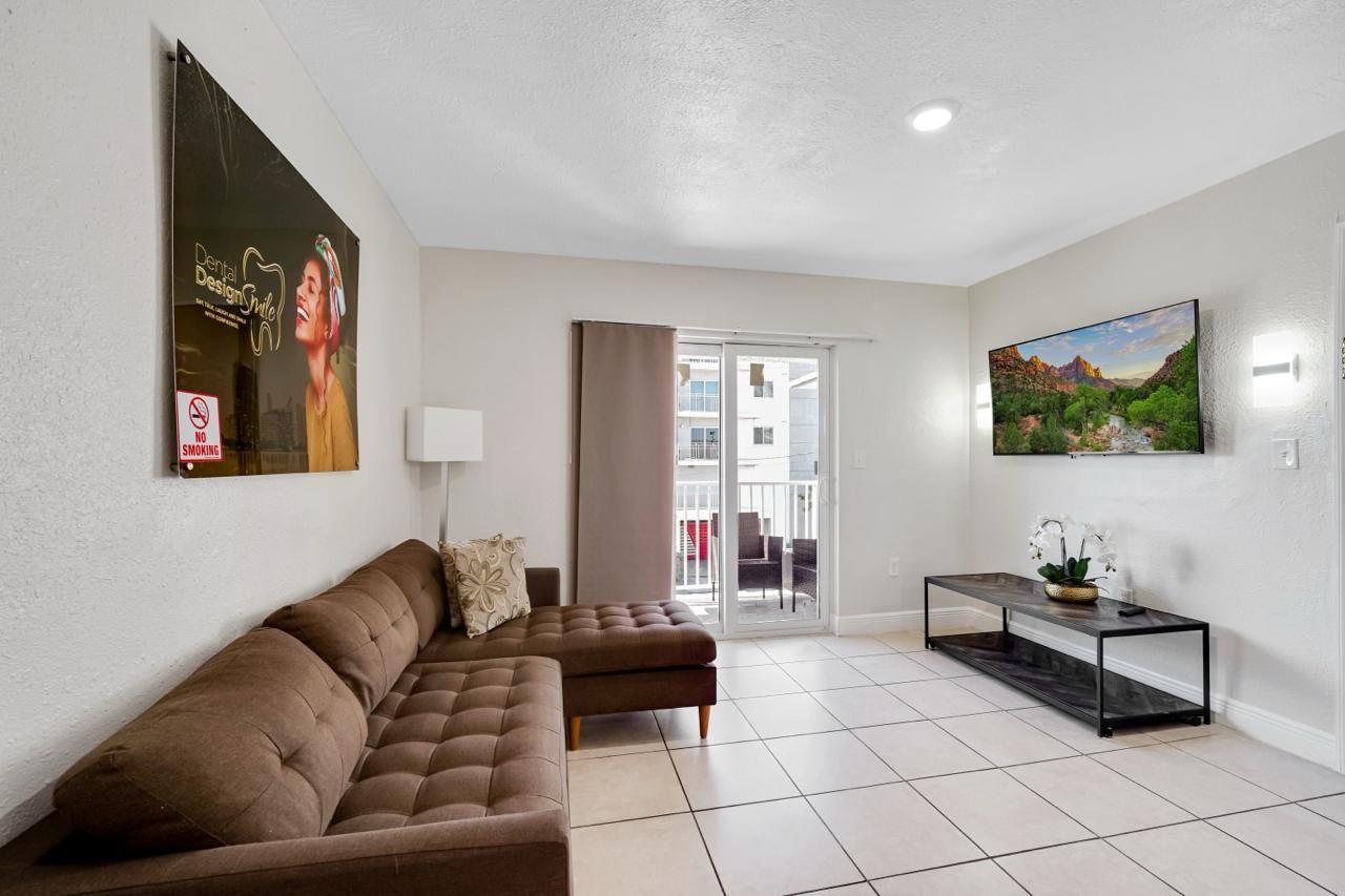 A modern living room with a brown sectional sofa, tiled floor, a large wall-mounted TV, and a glass sliding door balcony.