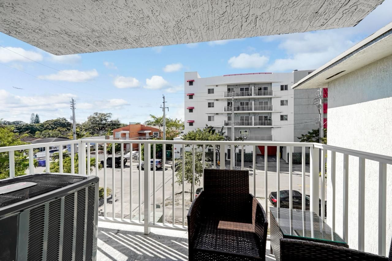 A balcony view with dark wicker chairs and an air conditioning unit overlooking a parking lot and a white apartment building.