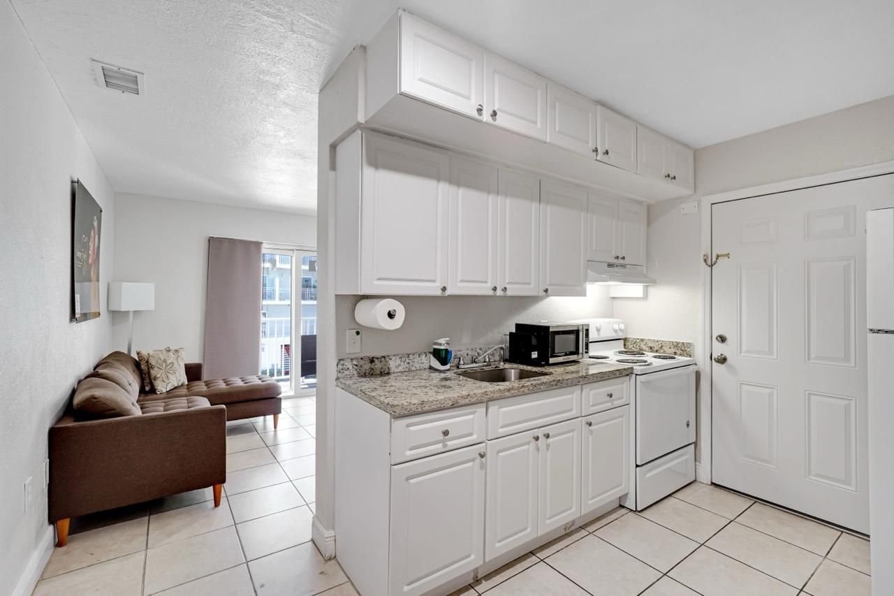 A compact kitchen with white cabinets, granite counters, and a white stove next to a living area with a brown sofa.