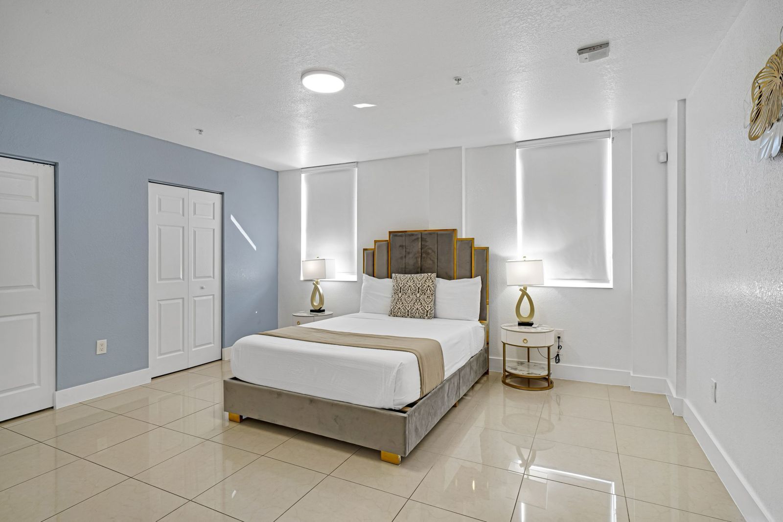 A bedroom with a queen-sized bed, grey tufted headboard, white bedding, and two lamps, set against a light blue wall.