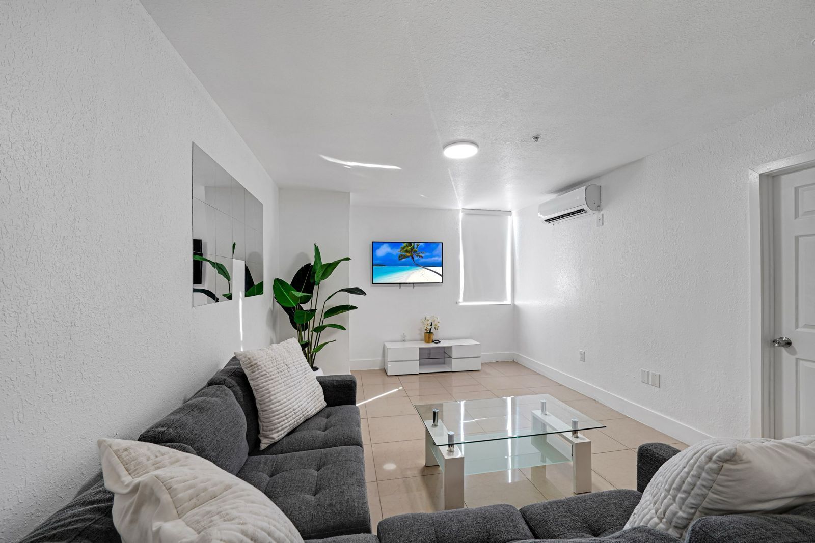 A bright living room features a dark gray sofa, a glass coffee table, a white media console, and a potted plant.