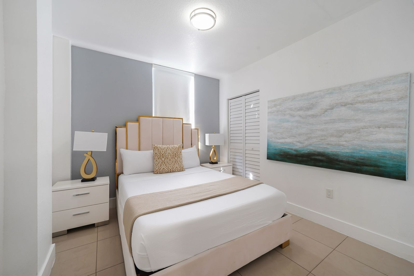 A bright bedroom with a cream tufted headboard, white bedding, gray accent wall, and a large teal landscape painting.