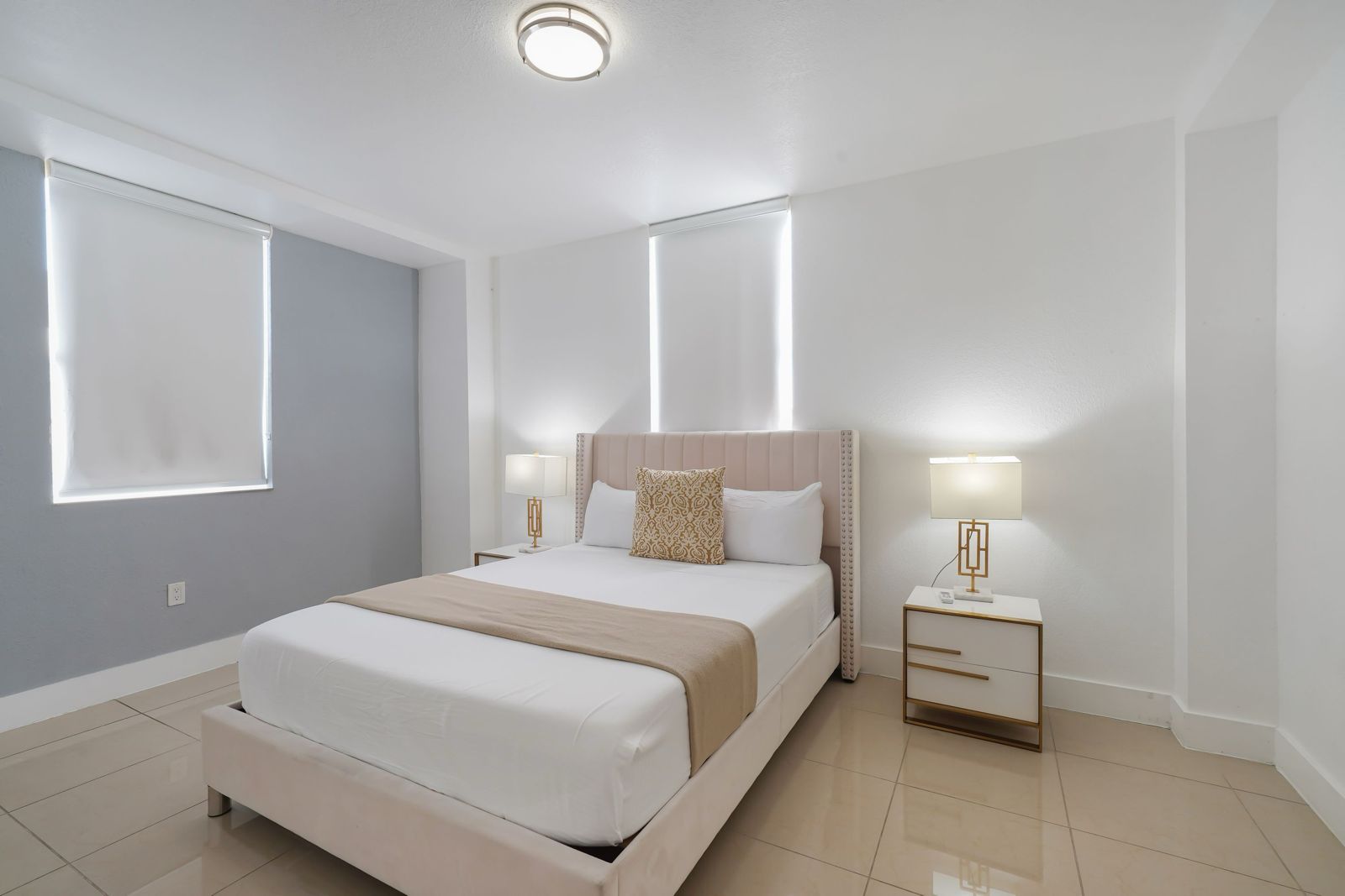 A modern bedroom with a beige upholstered bed, white linens, two white nightstands with gold lamps, and light tile floors.