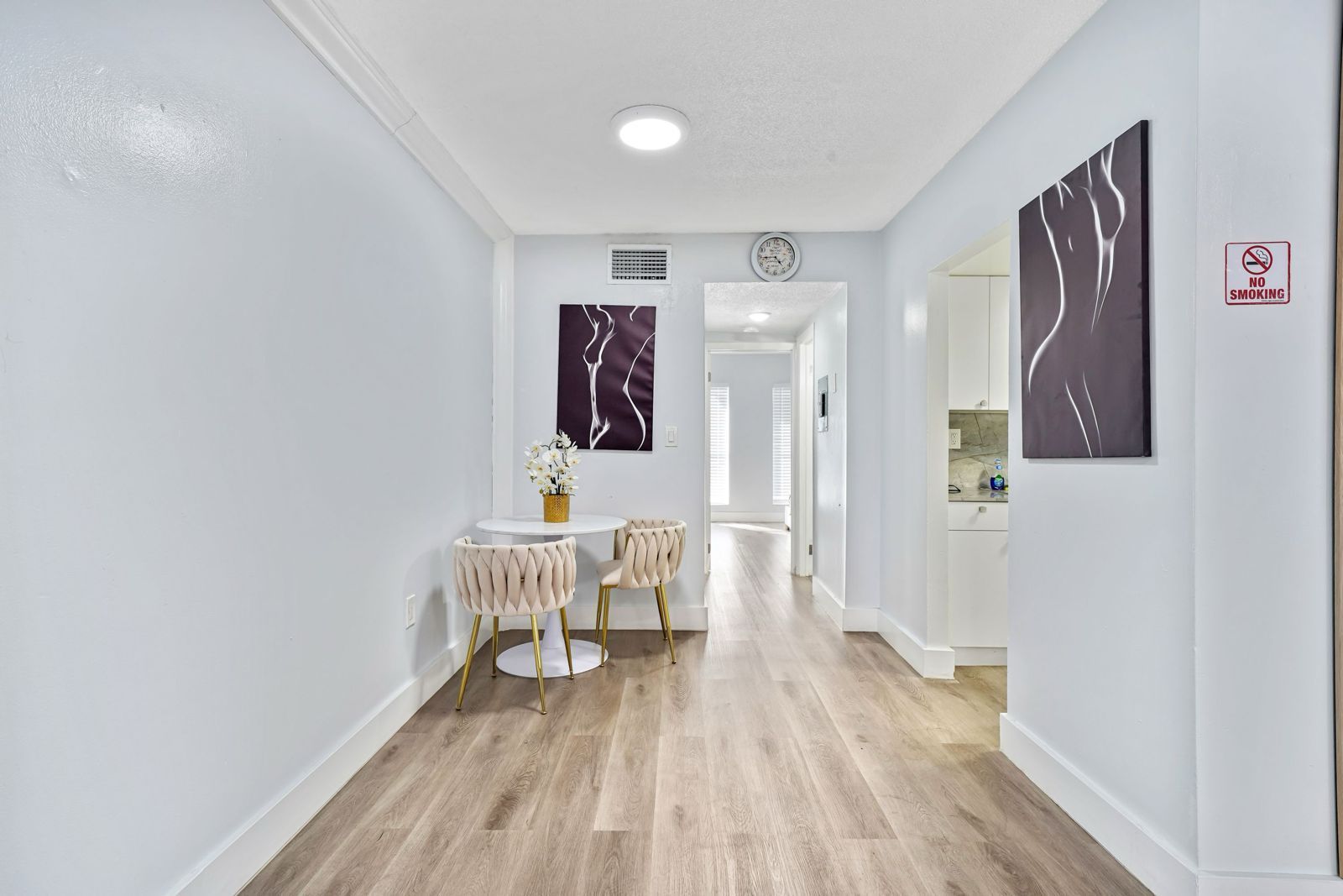 A hallway with light wood floors, a small white table with two chairs, two abstract wall prints, and an adjacent kitchen.