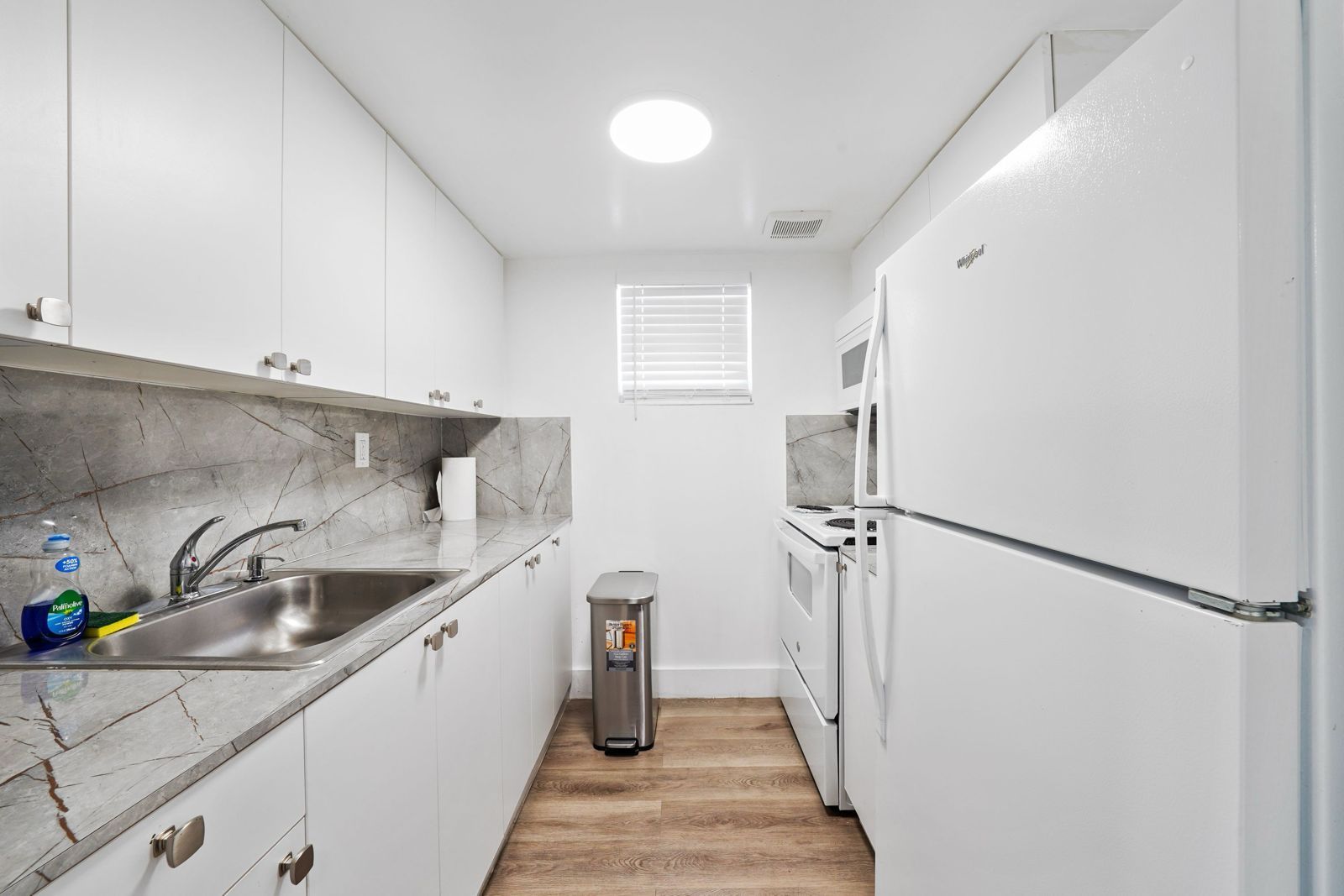 A galley-style kitchen with white cabinets, a stone backsplash, a sink, a stove, a white refrigerator, and wood flooring.