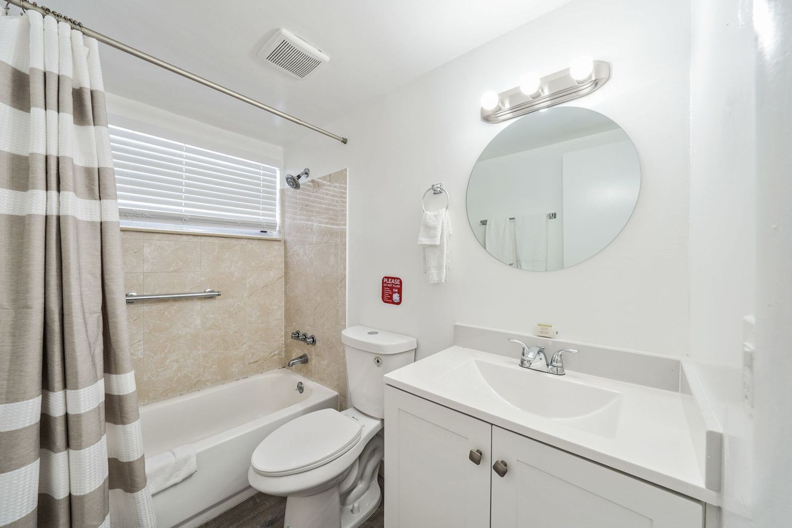 A bathroom with a white vanity, a round mirror, and a beige tiled bathtub area with a grey and white striped shower curtain.