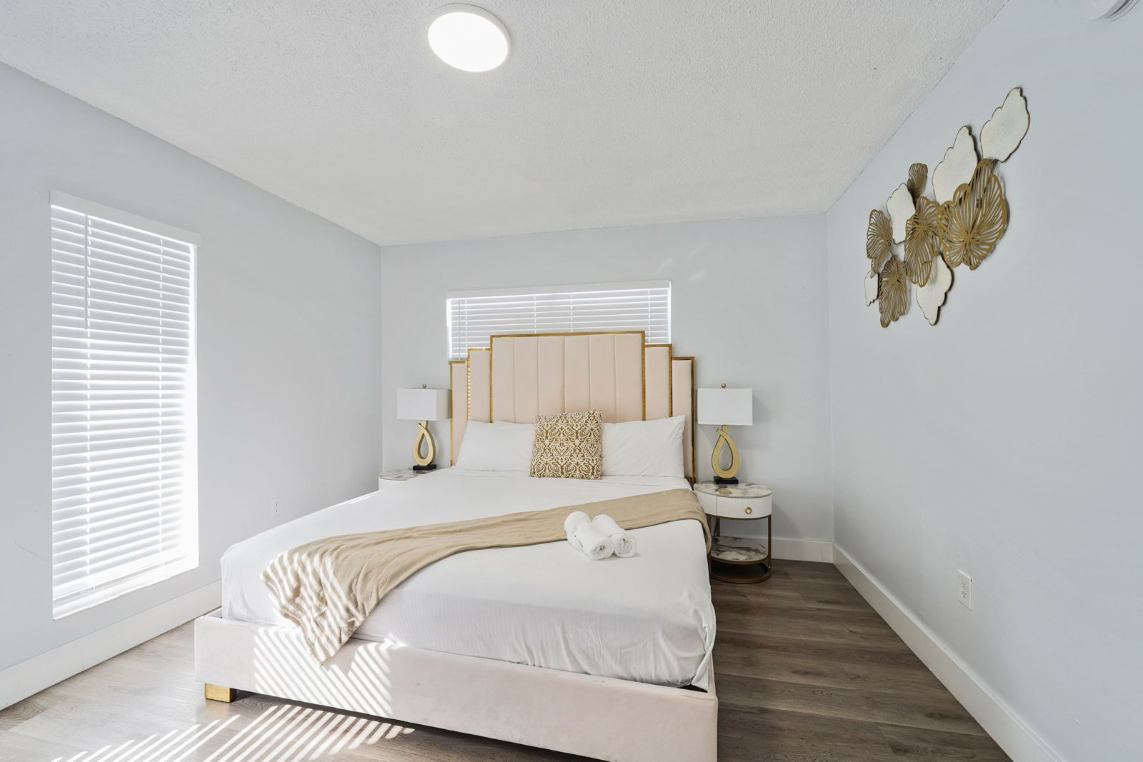 A bright bedroom with a cream tufted bed, white linens, bedside tables, and a metallic wall art piece.