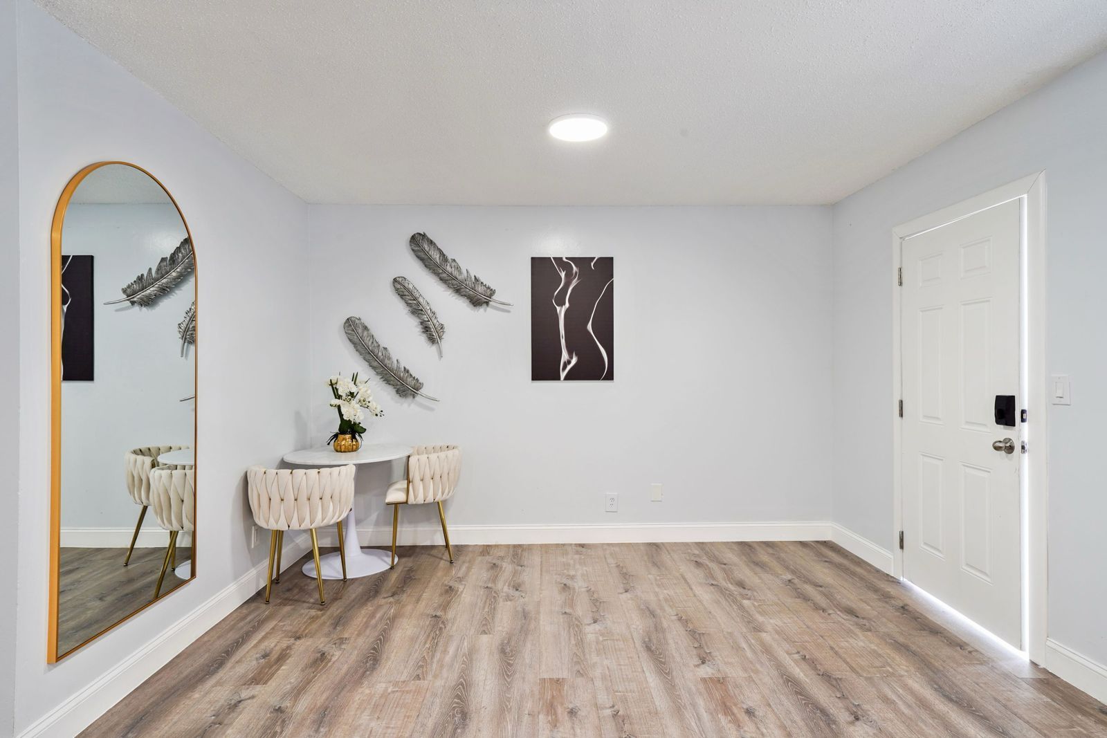 A minimalist room with light wood floors, a gold-arched floor mirror, a small white bistro table, and wall decorations.