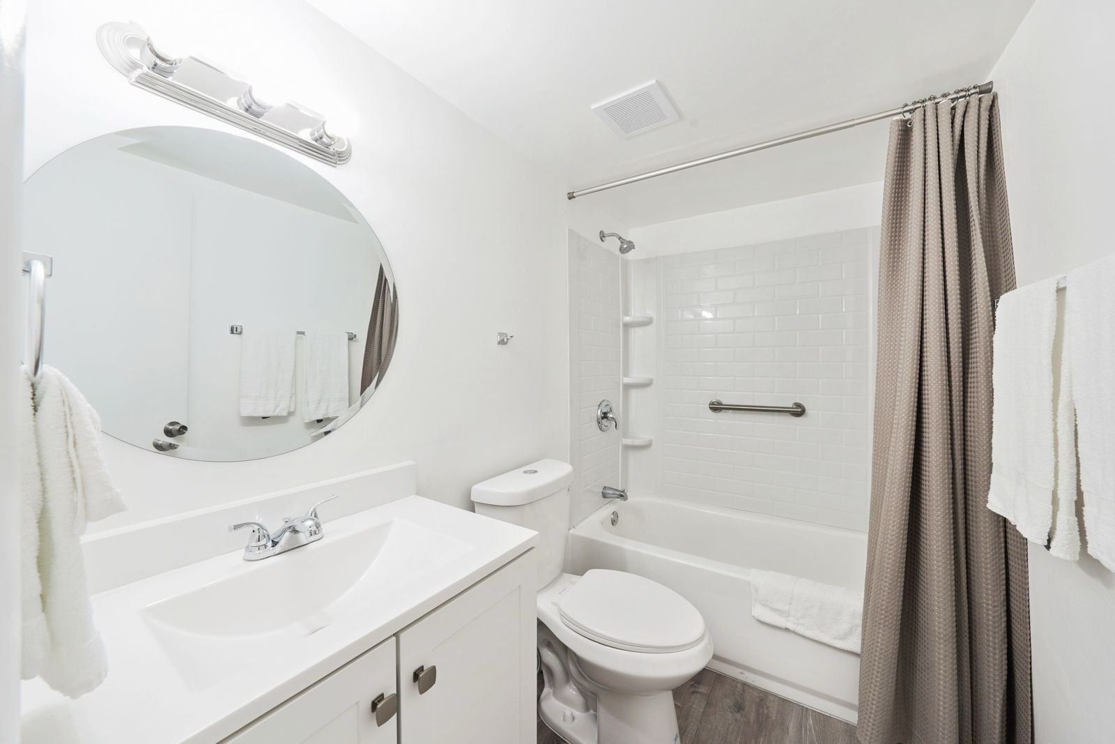 A white bathroom featuring a circular mirror, a vanity sink, a toilet, and a bathtub with a tan curtain.