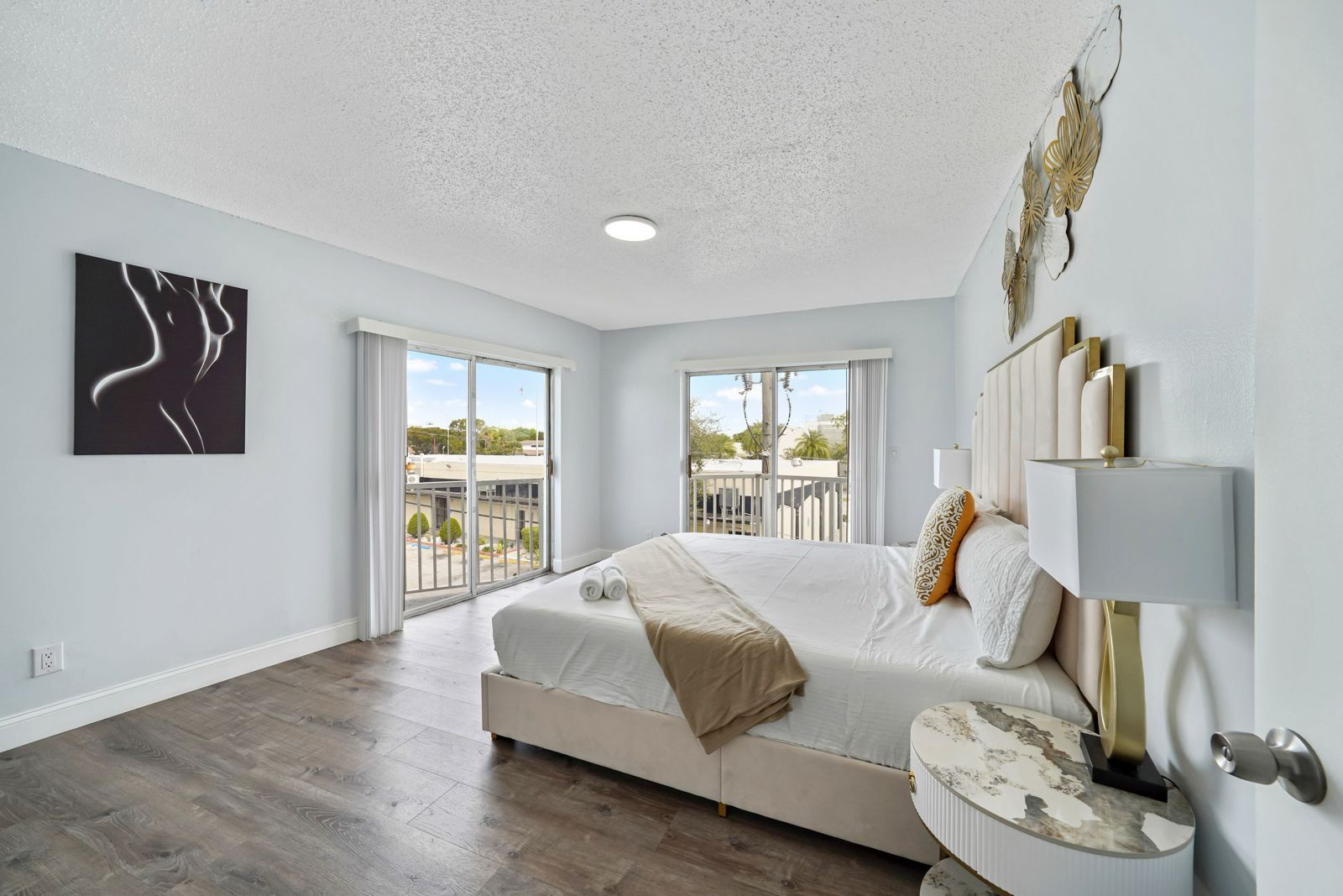 A bright, modern bedroom featuring a bed with white bedding, wood-look floors, a black wall art piece, and sliding doors.