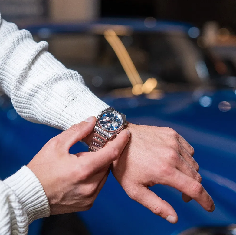 Uomo che indossa un orologio da polso argentato con il quadrante blu e tiene la mano nella tasca dei jeans.
