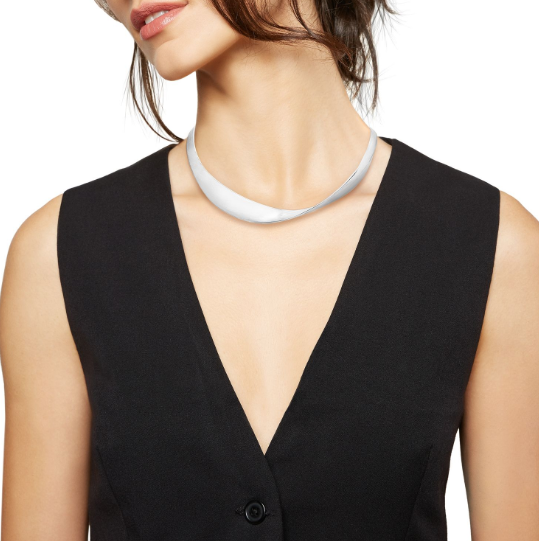 Donna che indossa un top bianco con le spalle scoperte, una collana d'argento con una pietra scura, e guarda la telecamera.