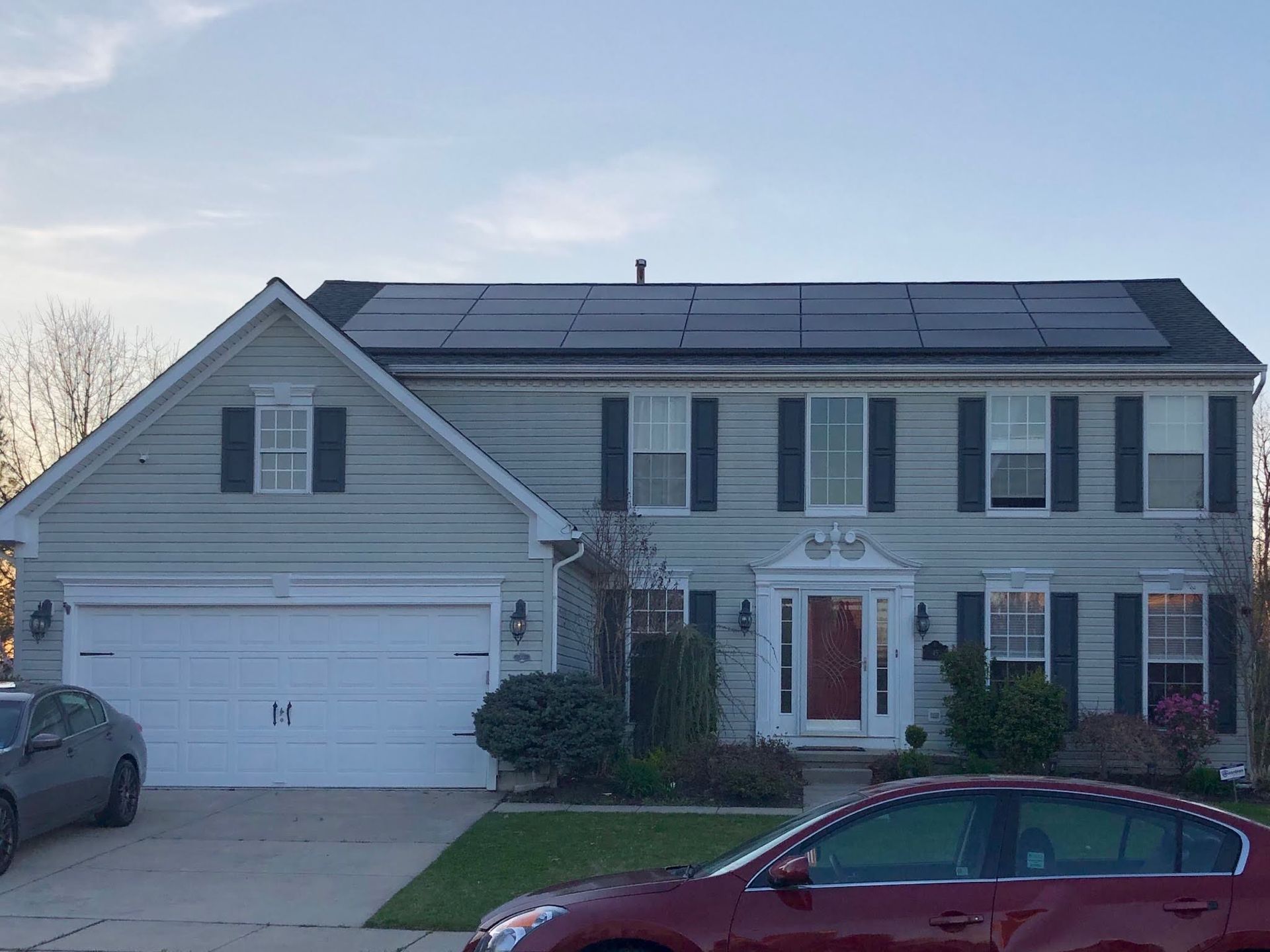 A red car is parked in front of a house with solar panels on the roof.
