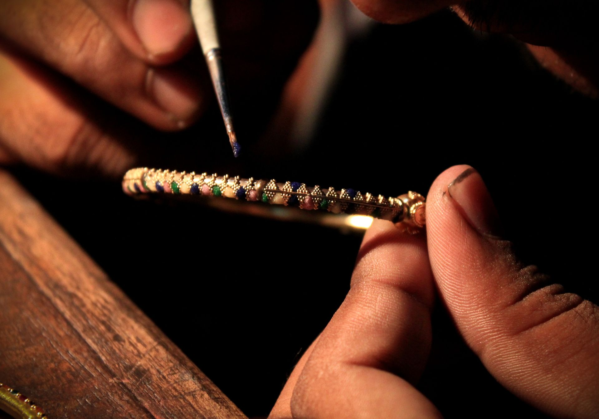 Mani che realizzano un braccialetto d'oro, applicando gemme colorate con uno strumento. Incastonatura scura.