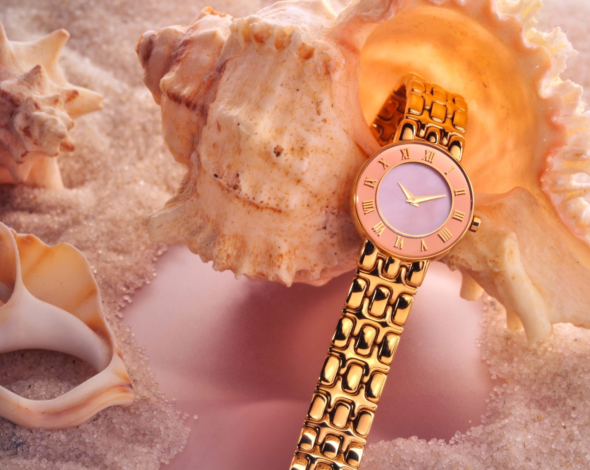 Orologio da polso in oro con quadrante rosa all'interno di una conchiglia, appoggiato sulla sabbia insieme ad altre conchiglie.