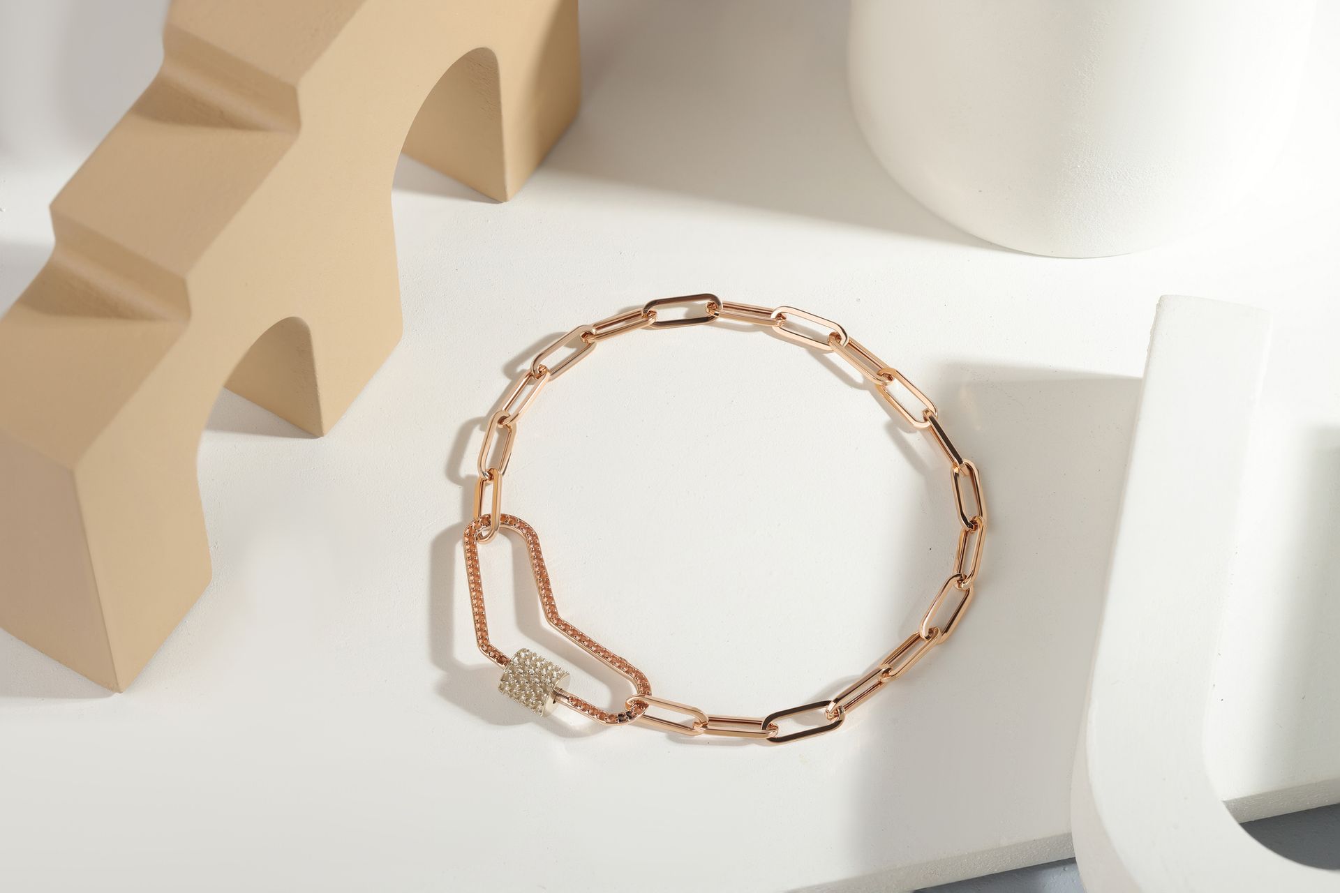 Bracciale a catena in oro rosa su superficie bianca con forme artistiche dai toni neutri.