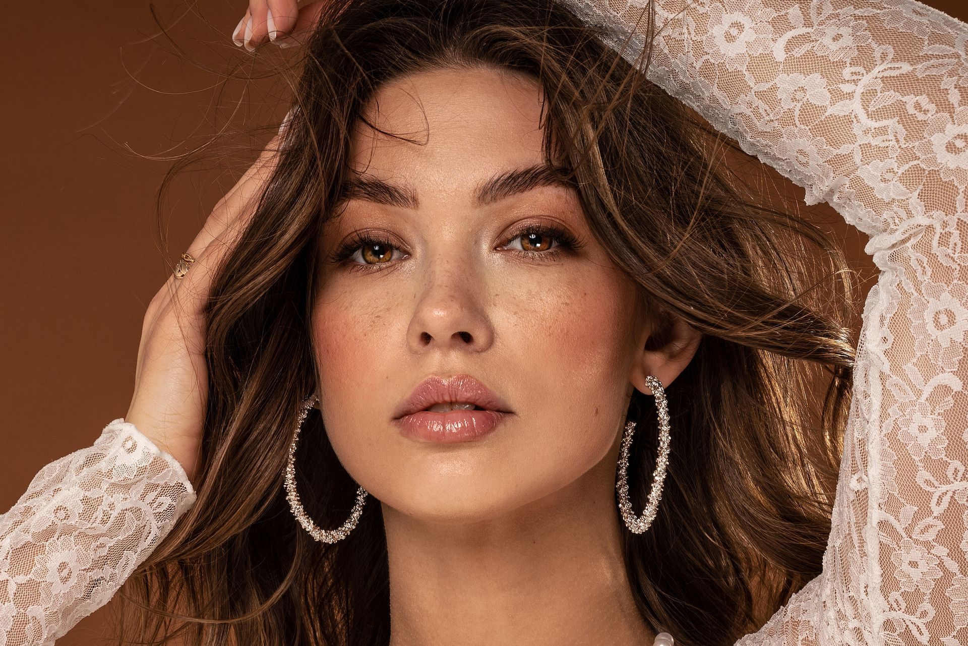 Donna con orecchini a cerchio, mani nei capelli, indossa un top di pizzo bianco, rivolta in avanti.