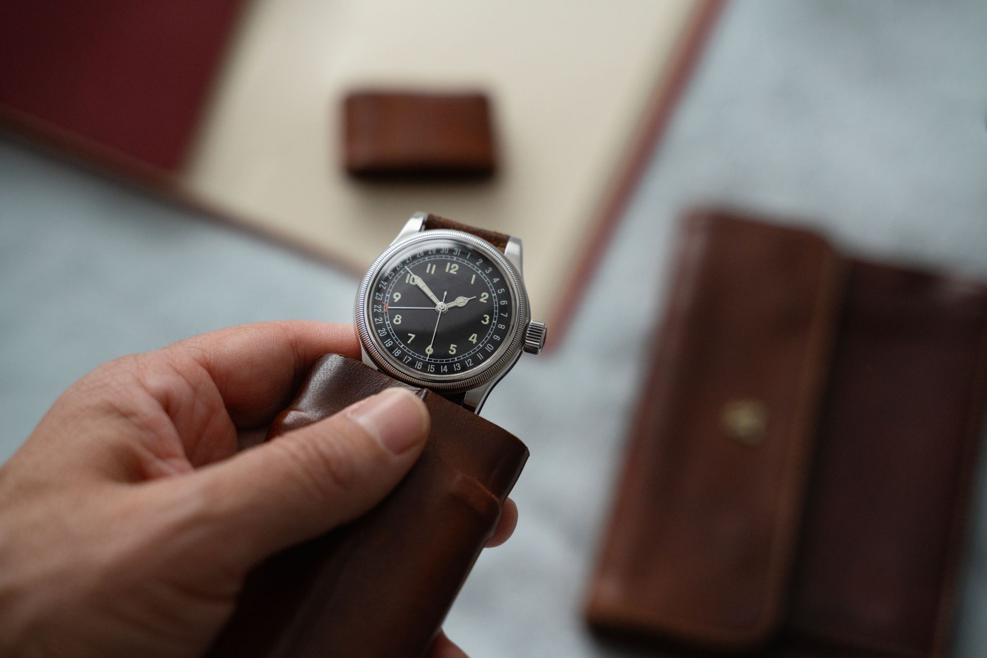 Una mano regge un orologio da polso argentato con il quadrante nero, custodito in una custodia di pelle marrone; sullo sfondo si vedono altri oggetti in pelle.