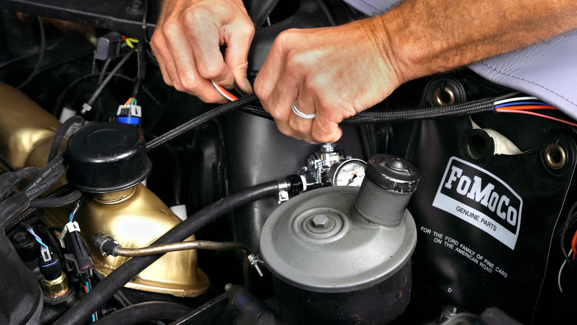 Hands working on car engine, wires. Black, gray, and gold components in engine bay.