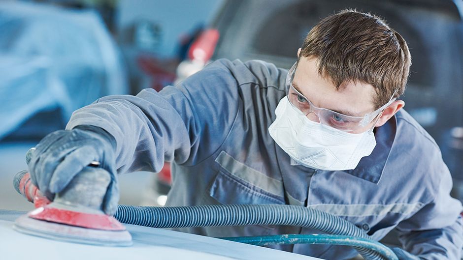 A person wearing protective gear sands a car's surface in an auto shop.