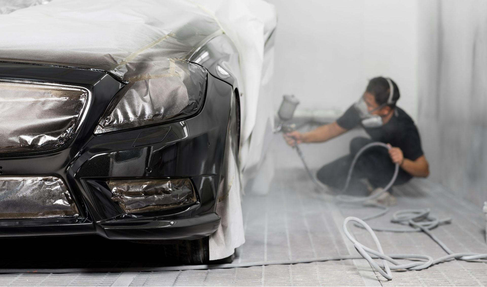 Car being painted black in a garage; a person in headphones sprays paint on the front.