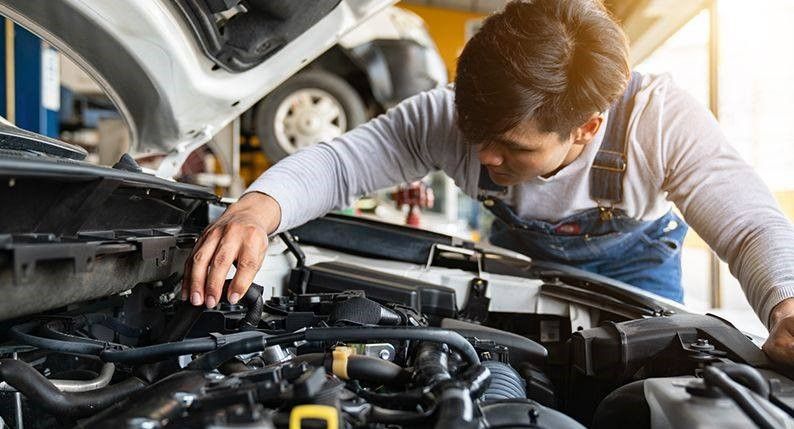 Mechanic examines car engine, hands on components. Garage setting, wearing overalls.
