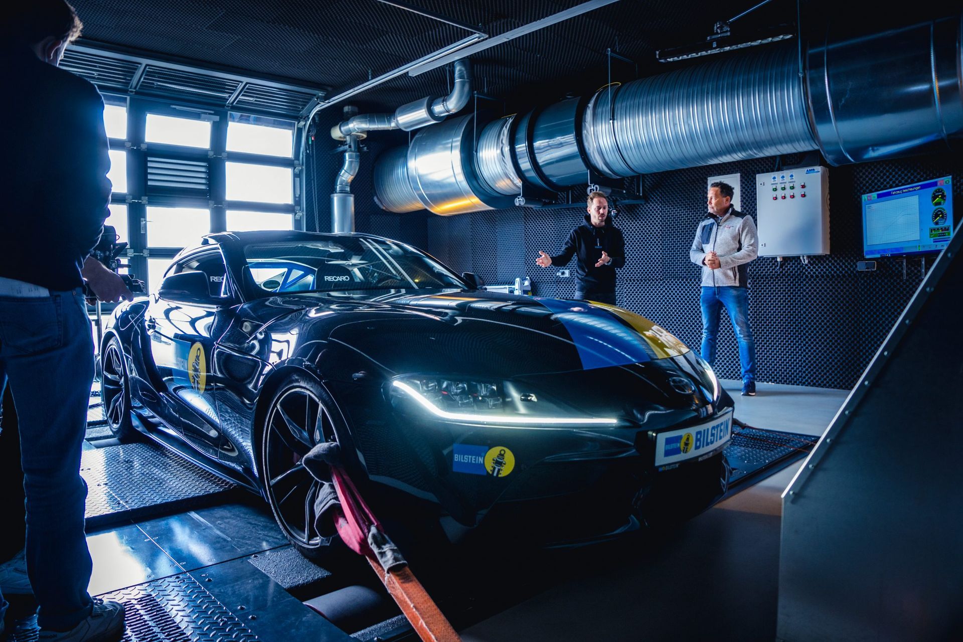 Car on a dyno in a lab. Two people observe. Dark green car with blue stripe, inside a building.