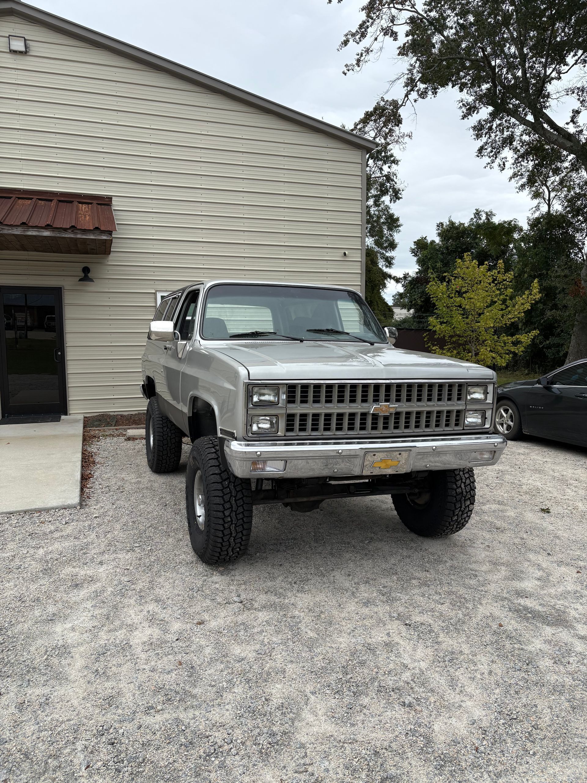 Silver lifted Chevy Blazer parked in front of a building with gravel ground.