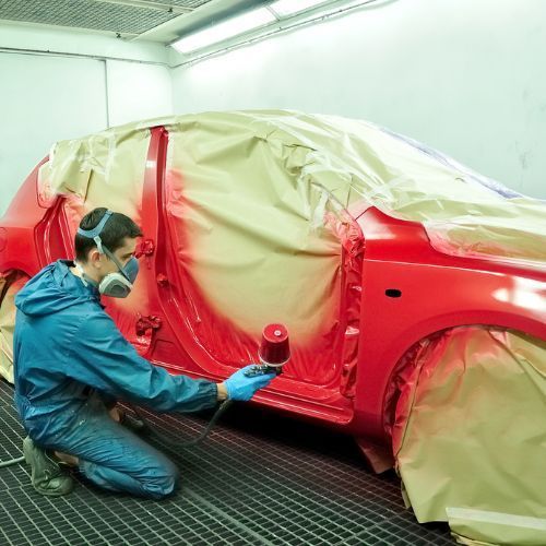 A person painting a red car in a body shop, wearing protective gear.