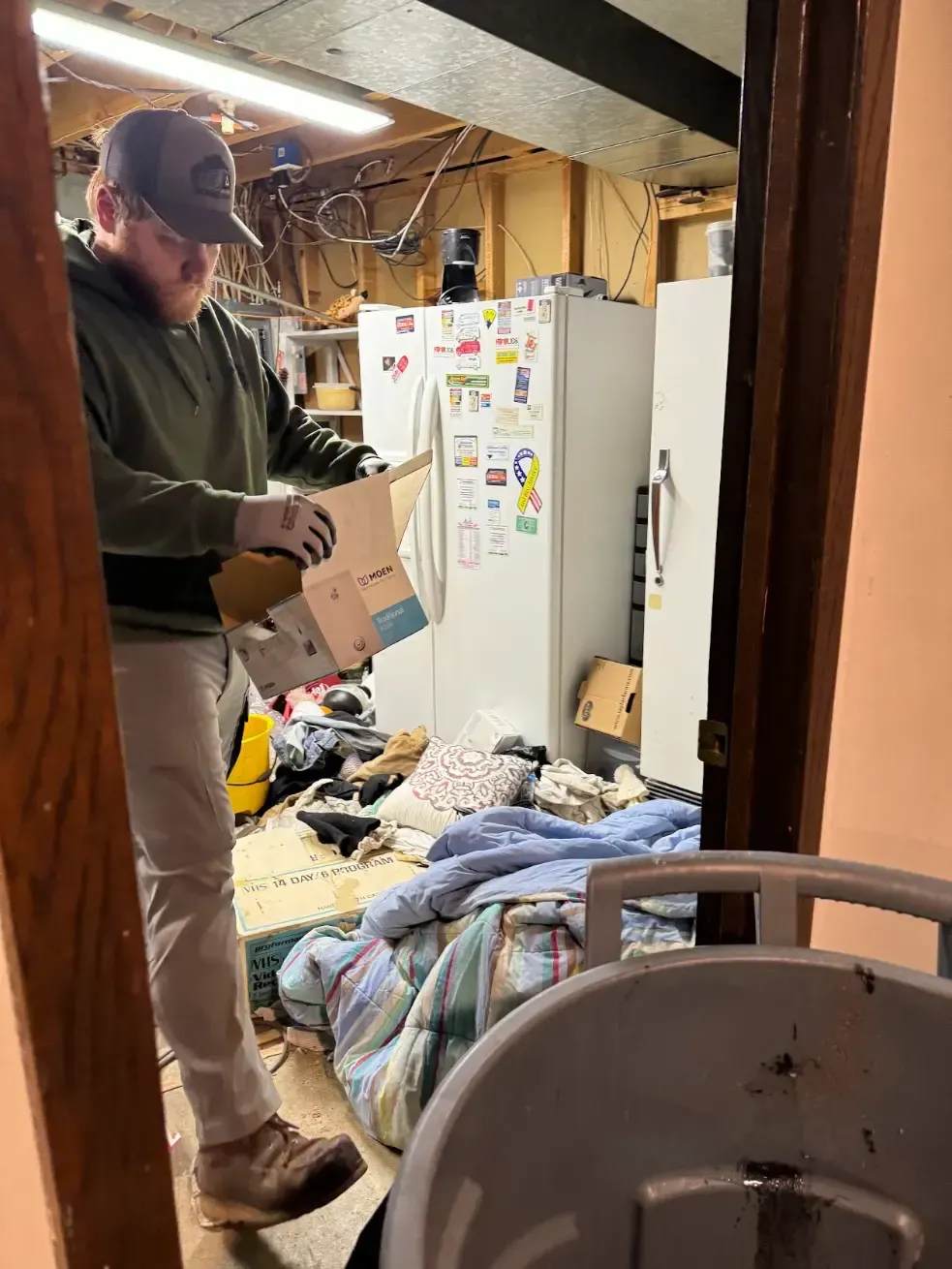 A man is standing in a messy room holding a box.